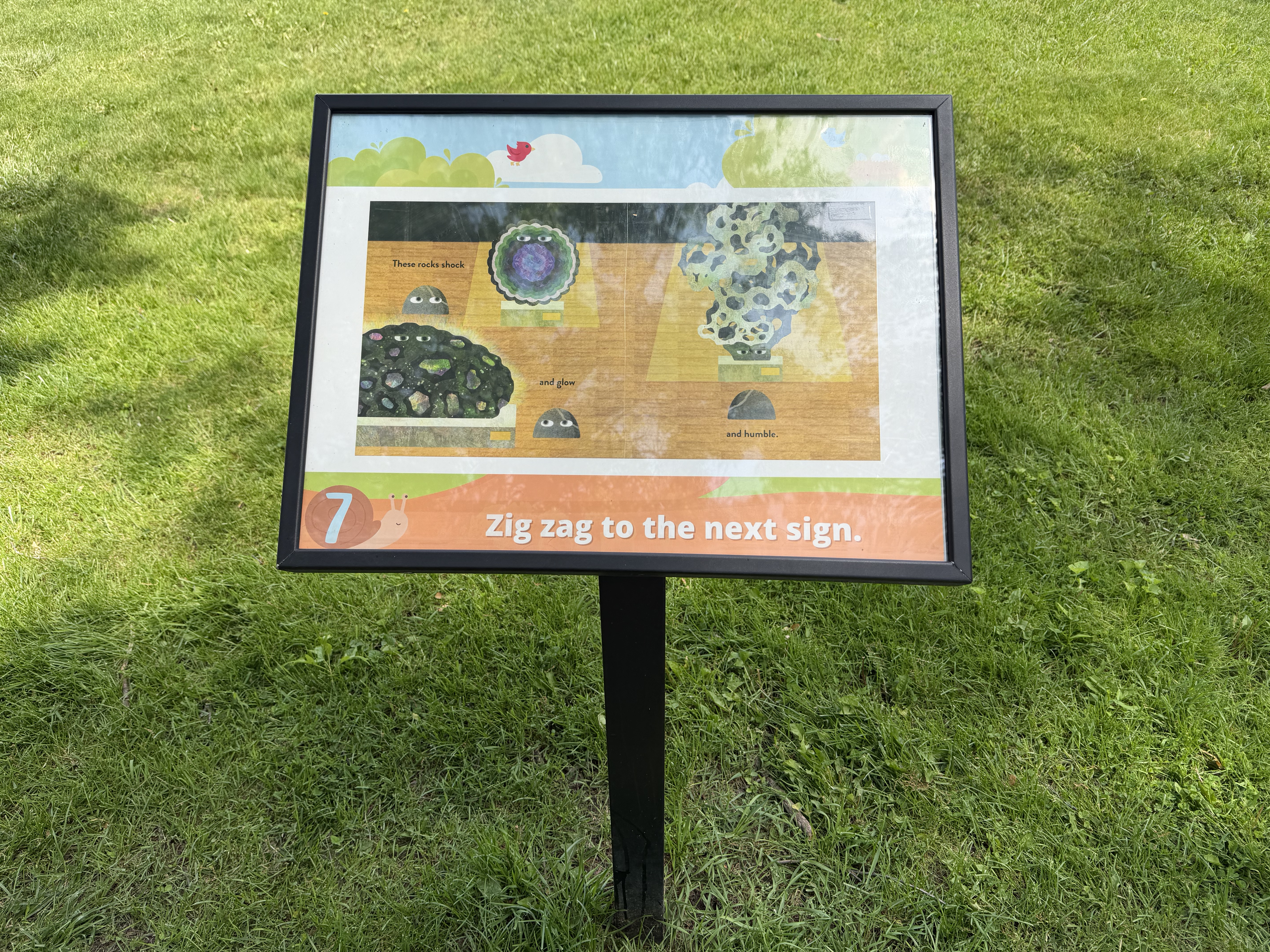 Close up of sign 7 with a book page in the middle. At the bottom of the sign is the action phrase "zig zag to the next sign". The background is green grass.